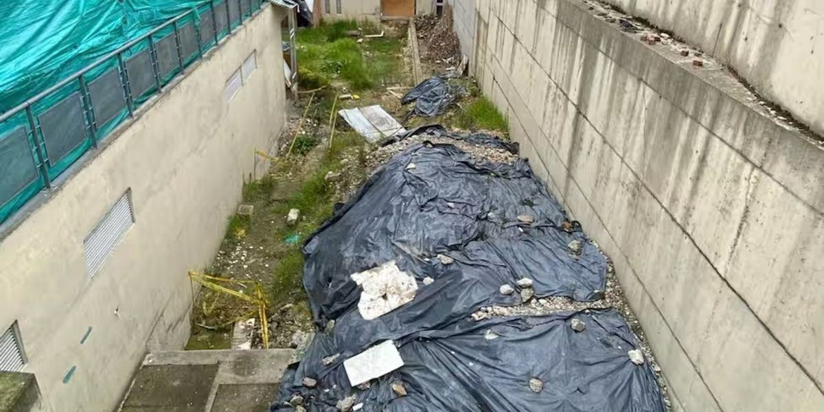 Canal entre dos muros de concreto con lonas negras sobre el suelo, escombros, vegetación dispersa y una cubierta verde a la izquierda.