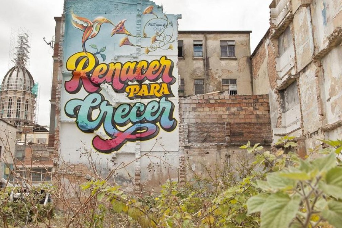 Solar baldío con maleza frente a edificios viejos. En una pared hay un mural colorido con colibríes y el texto grande: “Renacer para Crecer”.		