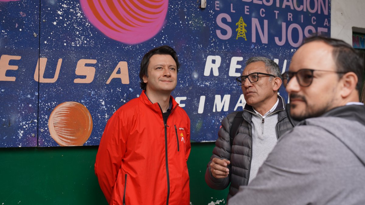 Tres hombres frente a un mural; uno viste chaqueta roja y otro sostiene un micrófono