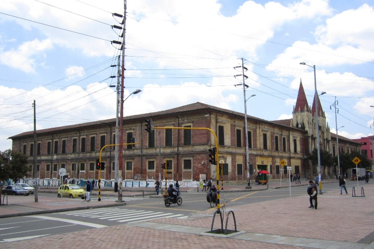 Edificio antiguo de ladrillo de dos pisos junto a una iglesia con torres puntiagudas, en esquina de avenida con semáforos; pasan peatones, motos, autos y hay carteles en la cerca.